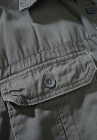 Detailní záběr na šedou látkovou kapsu s knoflíkovou klopou a viditelným šitím na oděvu.