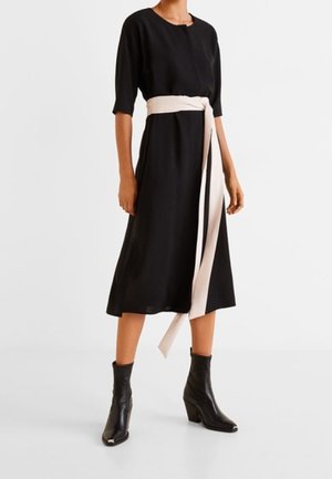 Henkilö käyttää mustaa midi-mittaista mekkoa, jossa on kyynärtaipeeseen ulottuvat hihat ja pitkä vaalea beige vyö sidottuna vyötärölle, yhdistettynä mustiin nilkkureihin.