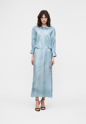 Kvinde med krøllet mørkt hår iført en lyseblå midi-kjole med lange ærmer, subtile blomsterprint og sorte åbne tå-hæle, stående på hvid baggrund.