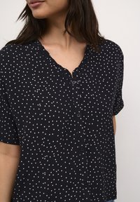 Blusa navy a maniche corte con pois bianchi, scollo a V con volant, chiusura a bottoni e texture del tessuto morbida.