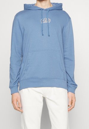 Persona con una sudadera con capucha azul de GAP con bolsillo delantero y cordones, combinada con pantalones blancos, mostrada desde la cabeza hasta las caderas.