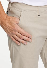 Pantaloni beige con un motivo testurizzato, dotati di una tasca frontale e una mano appoggiata sul tessuto, che mette in risalto il materiale e il design.