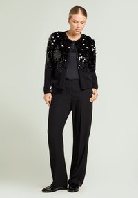 Cardigan nero con decorazioni di paillettes sulle spalle, indossato sopra una maglia nera e abbinato a pantaloni neri larghi e scarpe lucide.