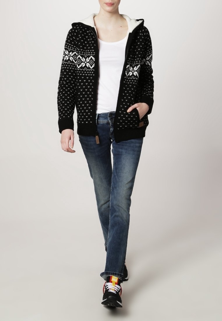 Personne marchant portant un sweat à capuche zippé à motif noir et blanc, un débardeur blanc, un jean bleu et des baskets noires avec des détails colorés.