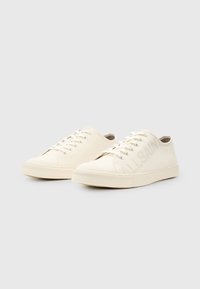 Sneakers bianchi in tela con sei fori, lacci piatti, suole in gomma testurizzata e un logo goffrato discreto sul lato.