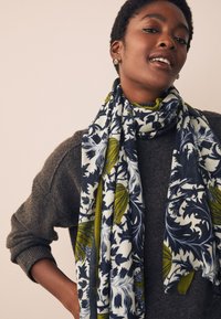 Écharpe à motifs avec design floral bleu marine, vert et crème, texture douce et bords frangés, enroulée autour d'un pull gris.