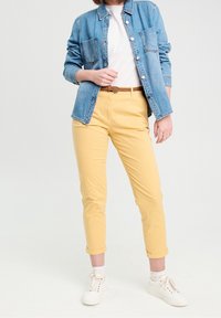Veste en jean avec deux poches avant, t-shirt blanc, pantalon jaune avec revers, et baskets blanches avec chaussettes à motifs. Tenue décontractée.