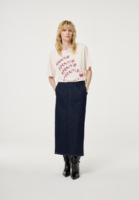 Crème t-shirt met roze tekst en een wijnglasafbeelding, gecombineerd met een donkerdenim maxi-rok. Model draagt zwarte, puntige enkellaarsjes.