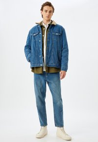 Giacca di jeans con colletto in pelliccia sintetica beige, lavaggio blu, tasche frontali, indossata sopra una camicia verde oliva. Jeans aderenti e sneakers beige.