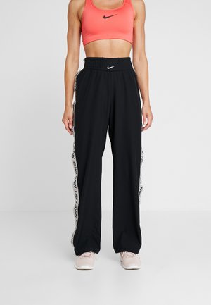 Kvinde iført sorte Nike træningsbukser med hvide sidestreger og en koralfarvet sports-bh, stående mod en ensfarvet lys baggrund.