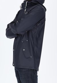 Veste imperméable marine avec capuche, dotée de quincaillerie argentée, de poches zippées et de poignets élastiques en un tissu lisse et texturé.