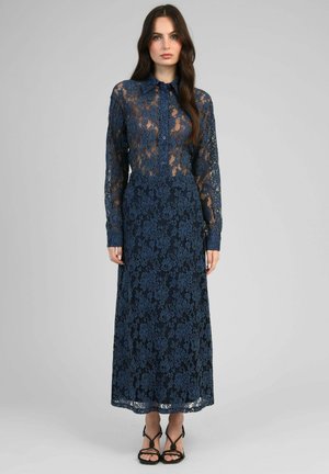 Femme portant une longue robe en dentelle bleu marine avec un col boutonné, des manches longues et des talons noirs à brides, debout devant un fond uni.