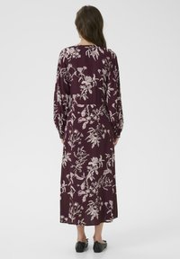 Eine Frau steht in einem langen, dunkelvioletten Kleid mit weißen Blumenmustern, das die Rückansicht zeigt, und trägt dazu schlichte schwarze Schuhe.