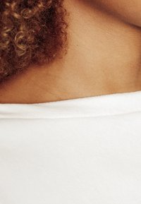 Top bianco scollato con un tessuto morbido e testurizzato. Capelli ricci in tonalità di marrone caldo incorniciano il décolleté, mettendo in risalto una pelle liscia.