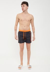 Pantaloni da nuoto neri con un elasticizzabile laccio arancione e la scritta "SUNDEK" stampata in arancione sul lato sinistro. Materiale liscio e leggero.