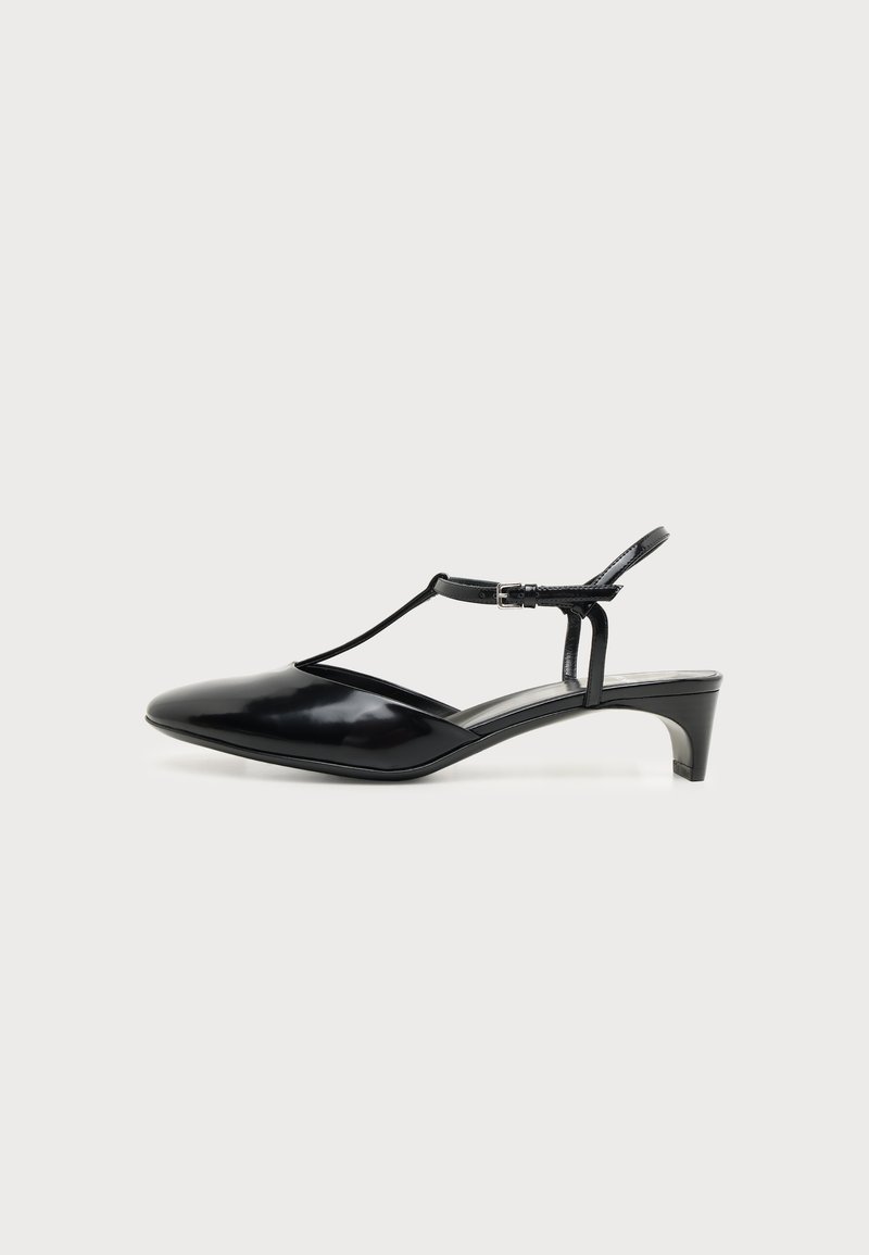 Chaussure noire en cuir verni à bout pointu et talon bas avec bride en T et boucle réglable, vue de profil sur fond blanc uni.