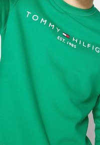 Felpa verde in cotone, con scollo rotondo e testo ricamato "TOMMY HILFIGER EST. 1985" con logo a striscia orizzontale.