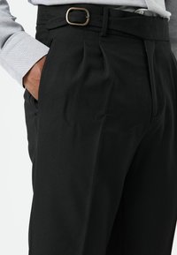 Zwarte op maat gemaakte broek met een gestructureerde stof, geplooid ontwerp en een metalen gespsriem. Twee zijzakken zijn zichtbaar.