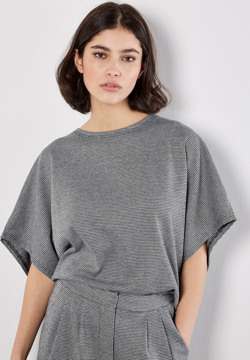 Joven mujer con cabello corto y oscuro viste un top holgado de patrón houndstooth en blanco y negro y pantalones plisados a juego, frente a una pared blanca.