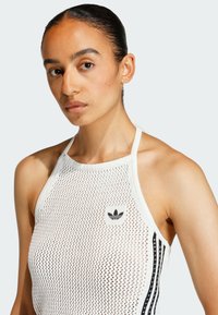 Bílé síťované tílko s kulatým výstřihem, černým logem Adidas a černobílými vertikálními pruhy na bocích. Texturovaný vzor látky.