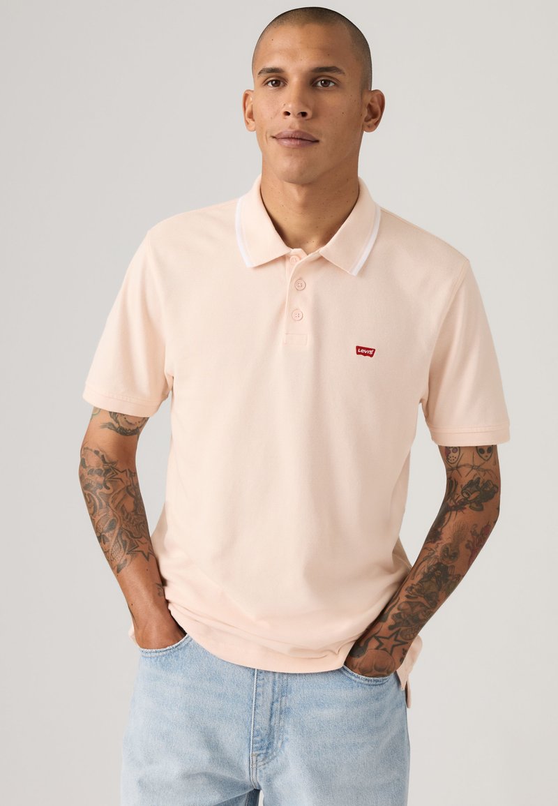 Homme tatoué portant un polo Levi's rose clair et un jean bleu clair, debout les mains dans les poches devant un fond uni.