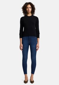 Pull noir à manches longues et ajusté, associé à un jean skinny bleu foncé et des ballerines en imprimé léopard. Le mannequin se tient devant un fond uni.