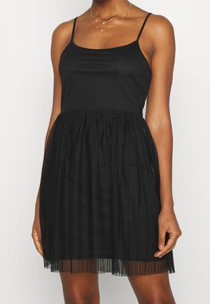 Vrouw draagt een mouwloze zwarte jurk met spaghettibandjes en een geplooide, semi-transparante rok, geaccentueerd met gelaagde gouden kettingen.