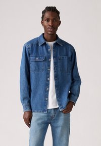 Chemise en denim bleu avec fermeture à boutons, deux poches poitrine, manches longues et coupe droite, portée sur un t-shirt blanc et associée à un jean clair.