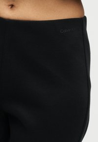 Κοντινή λήψη της μέσης ενός ατόμου που φοράει μαύρο παντελόνι Calvin Klein με ορατό λογότυπο κοντά στη μέση και εκτεθειμένο δέρμα πάνω από τη μέση.