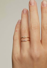 Anillos de doble banda de oro con un grupo de pequeñas piedras preciosas claras engastadas en la parte frontal. El fondo es neutro y la mano muestra una textura natural de la piel.