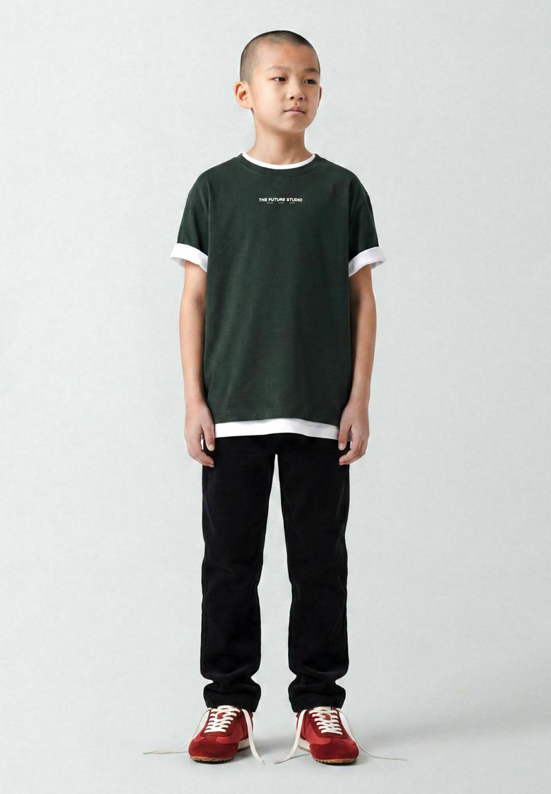 Garçon aux cheveux courts portant un T-shirt superposé vert foncé, un pantalon noir et des baskets rouges aux lacets défaits, debout devant un fond gris clair.