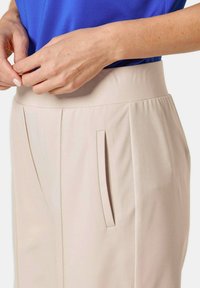 Beige Hosen mit hohem Bund, ausgestattet mit Messerfalten und seitlichen Taschen, kombiniert mit einem blauen Oberteil. Glatte Stofftextur, taillierte Passform.