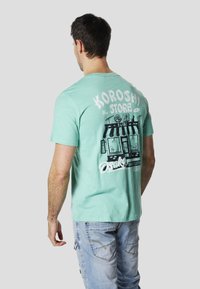 Camiseta de manga corta color verde menta con un estampado gráfico de una tienda en la espalda. Tejido suave y ligero con un corte relajado.