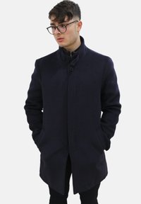 Cappotto blu scuro con collo alto, chiusura a bottoni e tasche laterali, realizzato in tessuto testurizzato con un taglio su misura.