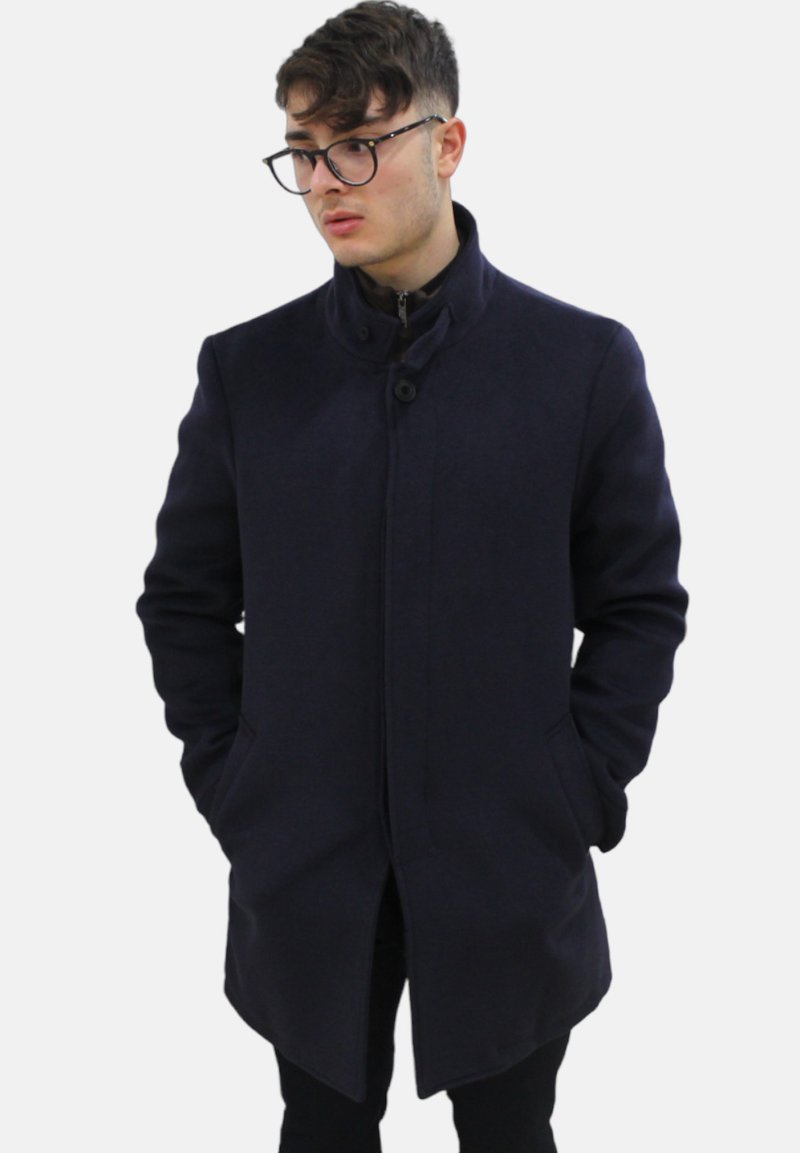 Cappotto blu scuro con collo alto, chiusura a bottoni e tasche laterali, realizzato in tessuto testurizzato con un taglio su misura.