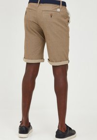 Brown shorts made of textured fabric with rolled cuffs, featuring a back pocket and small logo tag. Worn with black sneakers.