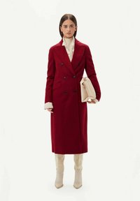 Röd ull dubbelknäppt kappa med två svarta knappdetaljer; krämfärgad höghalsad tröja under; håller i en beige kuvertväska; knähöga beige stövlar.