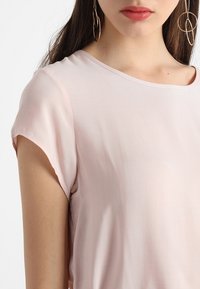 Ljusrosa blus som är gjord av mjukt, slätt tyg. Har en rundad halsringning och korta ärmar. Utmärkande drapering och minimala design detaljer.