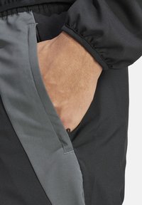 Schwarze Jacke mit grauen Taschendetails, ausgestattet mit einem Reißverschluss. Das Material scheint glatt und strukturiert zu sein, mit elastischen Bündchen an den Ärmeln.