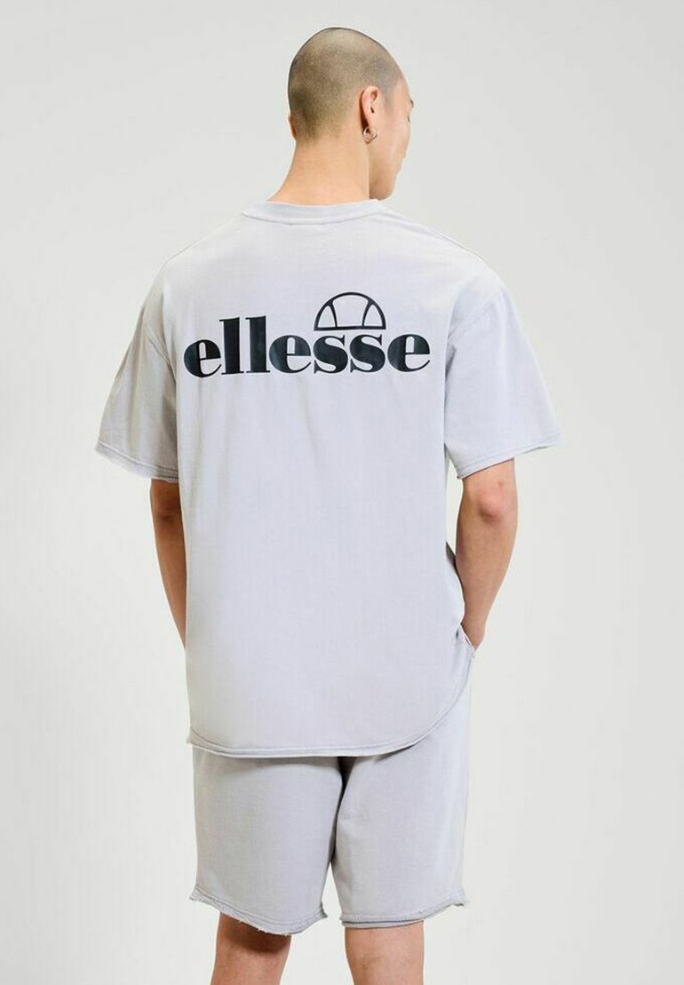 Homme avec la tête rasée portant un t-shirt ellesse gris clair et un short assorti, se tenant avec les mains dans les poches, tourné le dos au spectateur.