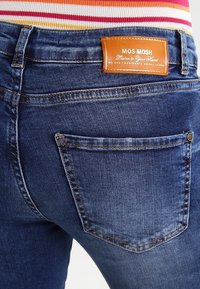 Donkerblauwe denim jeans met een enkele achterzak met een subtiele naaddetail en een leren merklabel in bruin, gestikt op de tailleband.
