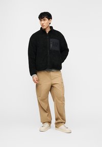 Νεαρός άνδρας που φοράει μαύρο φλις μπουφάν με τσέπη στο στήθος, μπεζ παντελόνι cargo και λευκά αθλητικά παπούτσια, στέκεται με το ένα χέρι στην τσέπη.