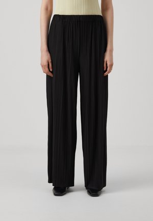 Femme portant un pantalon noir plissé taille haute à jambes larges associé à un haut sans manches côtelé de couleur claire et des chaussures noires.