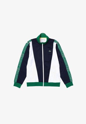Marineblaue, weiße und grüne Jacke mit Reißverschluss, grünen Bündchen und Kragen, Lacoste-Logo auf der Brust und wiederholtes Branding auf den Ärmeln.