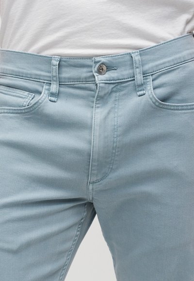 Homme portant un jean bleu clair et un t-shirt blanc, mise au point sur la zone de la taille avec bouton et poches visibles.
