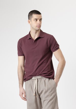 Hombre vistiendo una camisa polo color granate y pantalones beige con cordón, mirando hacia un lado contra un fondo claro y liso.