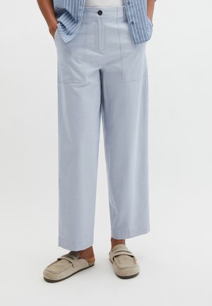 Jasnoniebieskie spodnie o szerokich nogawkach z przednimi naszywanymi kieszeniami, noszone z beżowymi wsuwanymi loafersami i niebiesko-białą paskowaną koszulą częściowo wpuszczoną do środka.