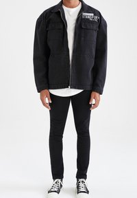 Musta farkkutakki, jossa on vetoketjullinen etuosa, kaksi rintataskua ja valkoinen teksti "STANDPOINT." Päällä valkoinen T-paita ja mustat slim-fit housut.
