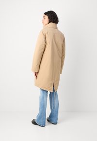 Kvinne som har på seg en beige knelange frakk med høy krage, lyseblå vide jeans og svarte sko, stående mot en enkel hvit bakgrunn.