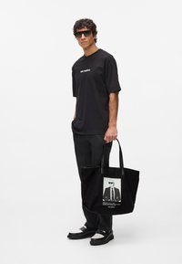 Crna torba s fotografijom figure koja nosi naočale, kožne remenčiće i mekanu teksturu. Model nosi crnu majicu i hlače.